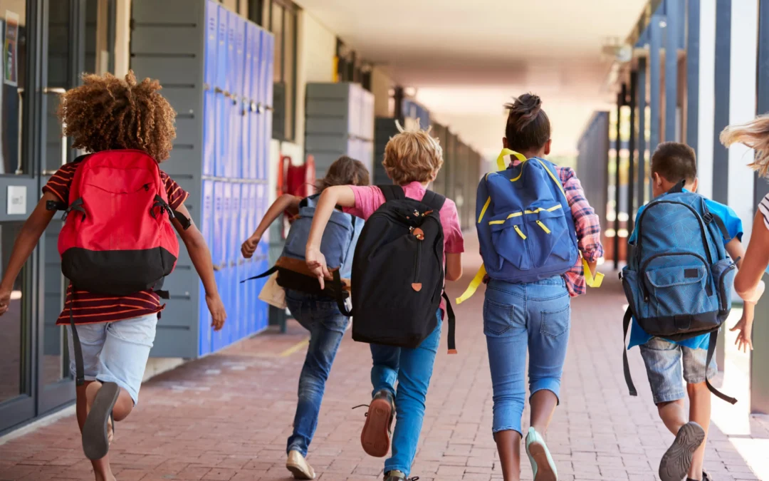 Group of children with backpacks running down a school hallway beside blue lockers on a sunny day.