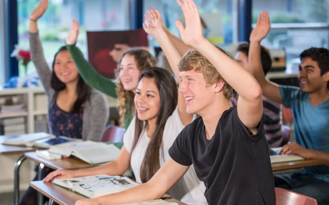 Group of students in a classroom raising hands and smiling during a lesson at desks with notebooks open on the desks.