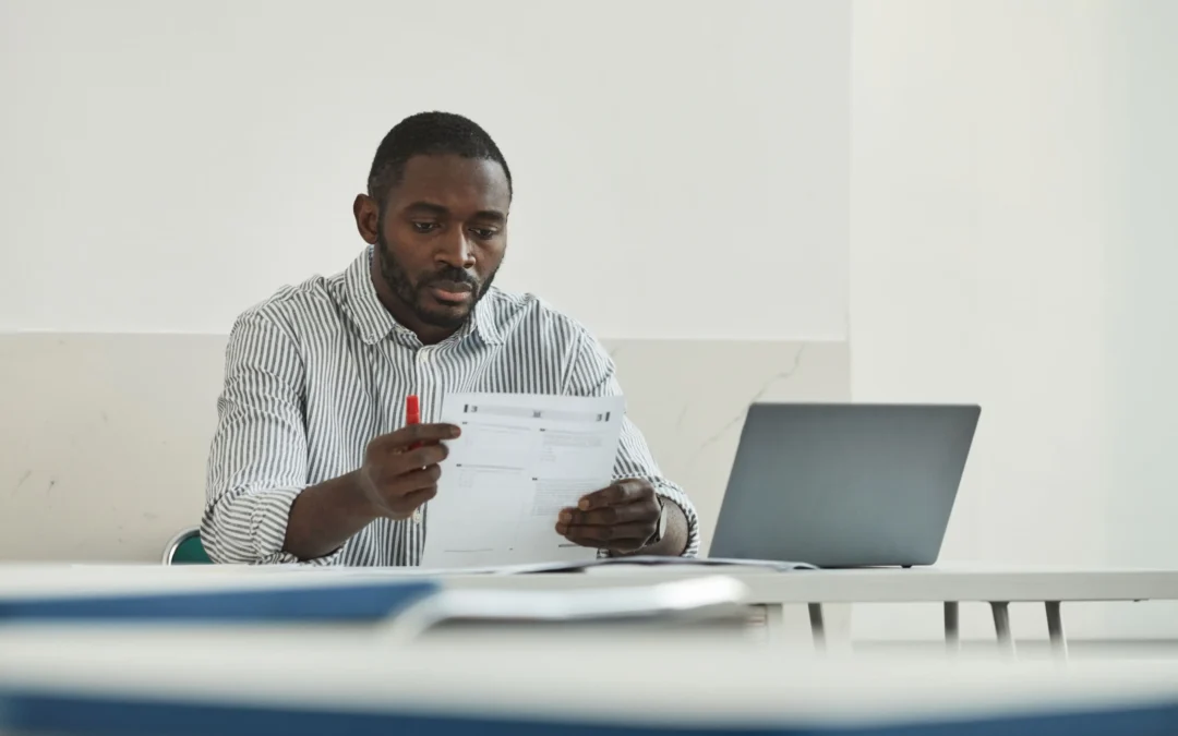 Man in striped shirt seated at a desk, reading a document with a red pen, a laptop open beside him.