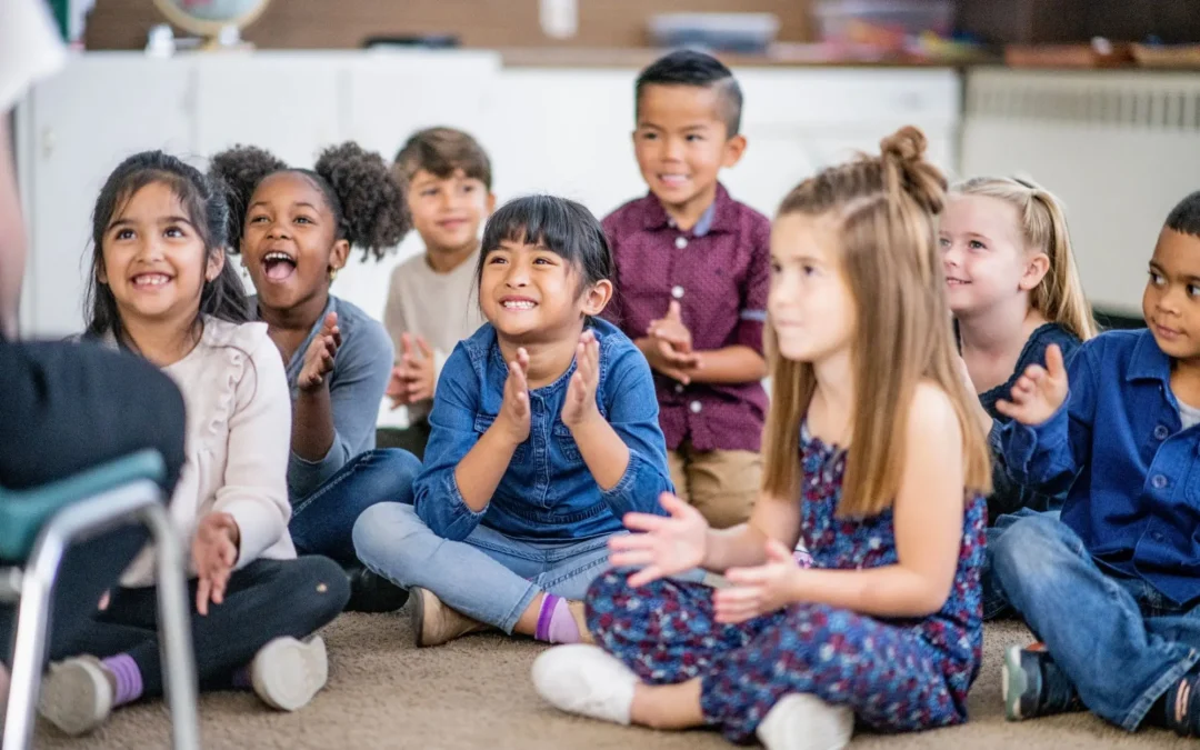Group of diverse children sitting on the classroom carpet, clapping and smiling.