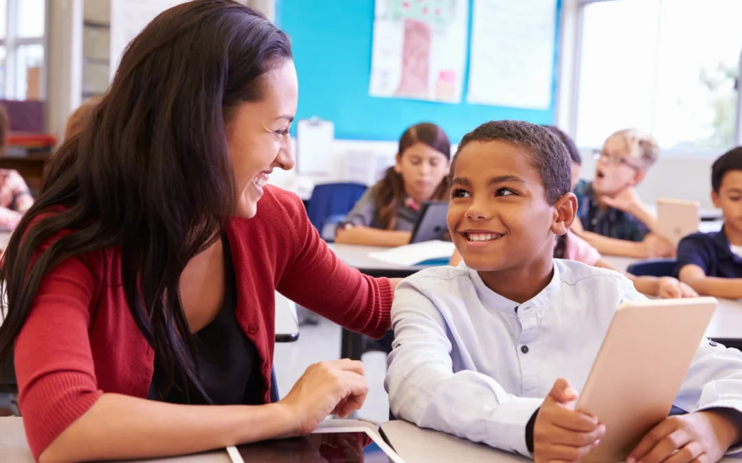 Teacher in a red cardigan smiles at a young student with a tablet in a bright classroom setting.